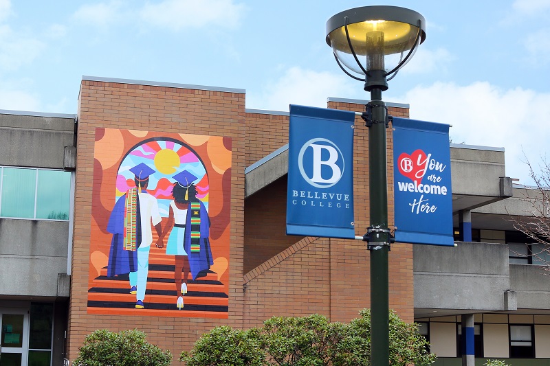 Image of wheatpaste mural Unity by Stat the Artist for Addressing Anti-Blackness Taskforce, shown beside a banner for Bellevue College. The mural depicts two of Black students in graduation regalia walking hand-in-hand up a flight of stairs toward a colorful horizon. The work is called 